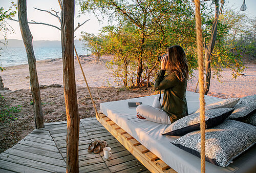 Eine Frau sitzt auf einer Holzveranda im Changa Safari Camp mit ihrer Kamera und macht ein Foto des Sees