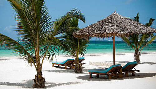 Sonnenliegen unter schattenspendendem Schirm auf weißem Sandstrand am türkisblauen Ozean zwischen Palmen