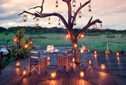 Ein Tisch und Stühle stehen auf einer Holzveranda, die um einen alten Baum gebaut wurde. Alles ist mit Öllampen dekoriert.