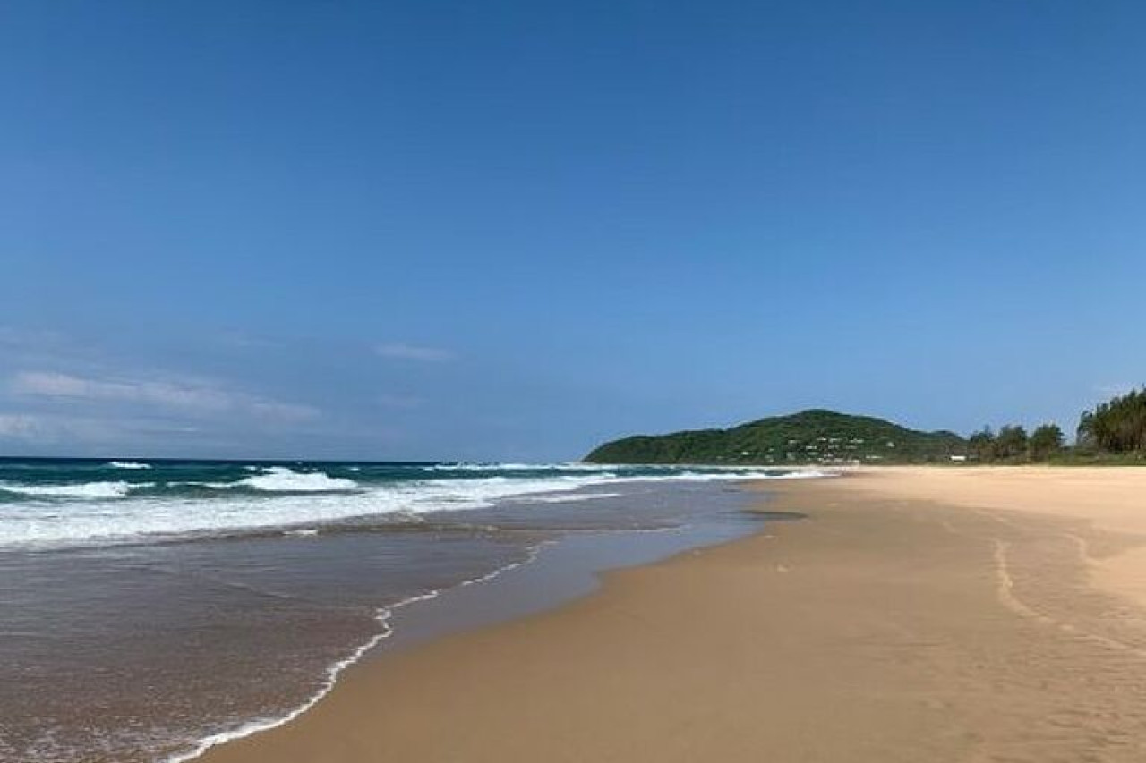 Weiter Sandstrand mit Wellen und grünen Hügeln.