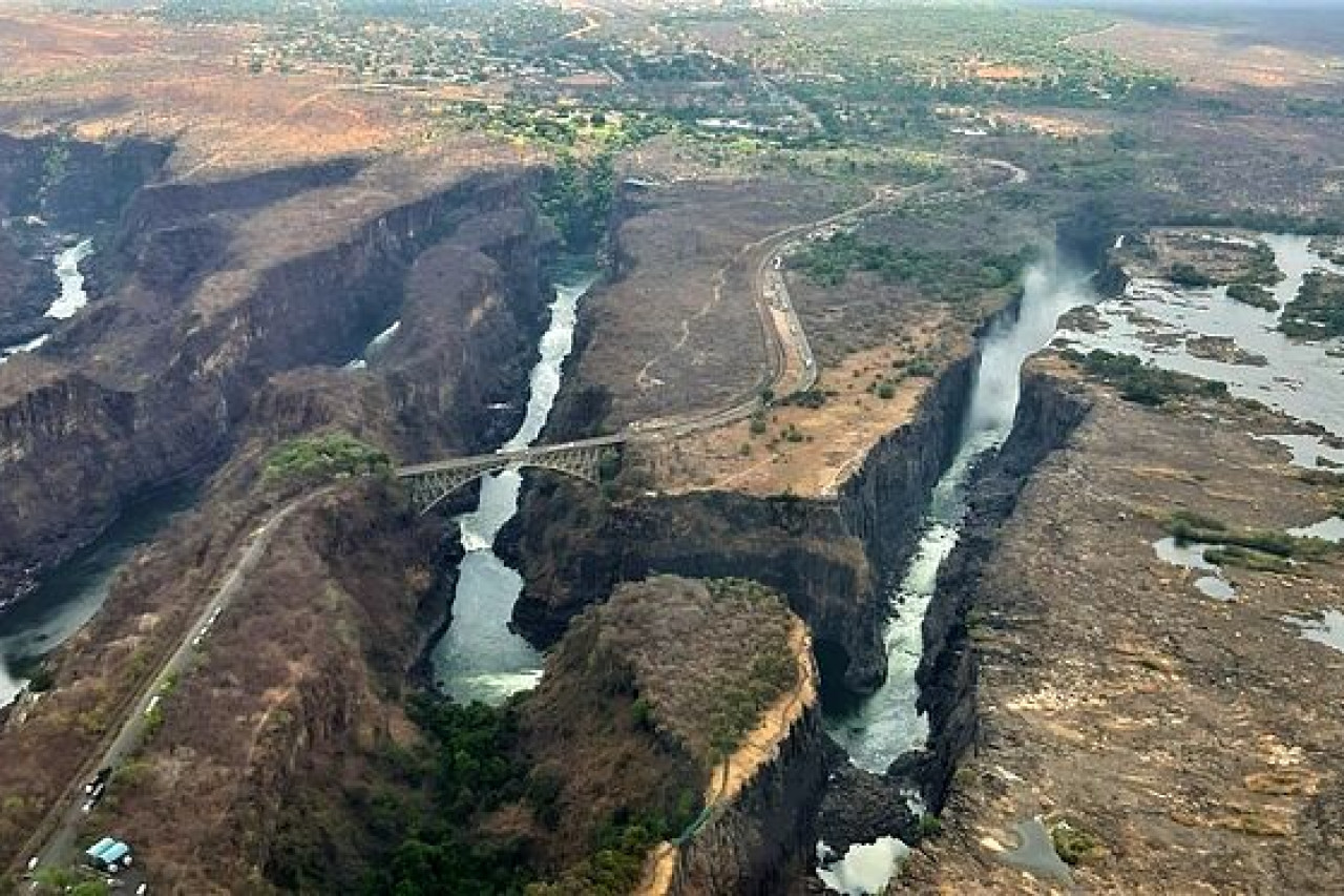 Luftaufnahme einer beeindruckenden Schlucht mit einem schmalen Fluss, der durch die tiefen Felsen fließt. In der Mitte der Szene ist eine Brücke sichtbar, die über den Fluss führt. Auf der rechten Seite sind Wasserfälle zu sehen, die in den Fluss münden. Die Umgebung ist von trockener Vegetation und einem sich schlängelnden Weg umgeben, während im Hintergrund die Landschaft sanft ansteigt.
