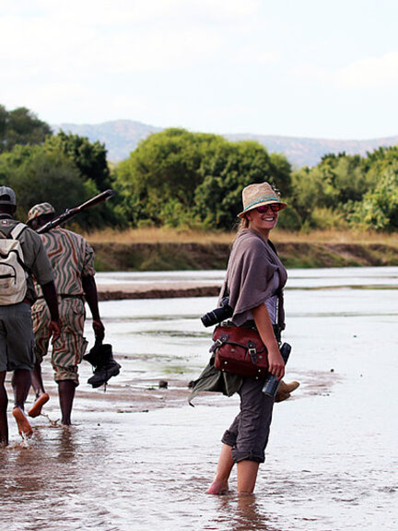 Frau auf einer Walking Safari in Afrika, begleitet von zwei Guides, die durch einen flachen Fluss laufen.