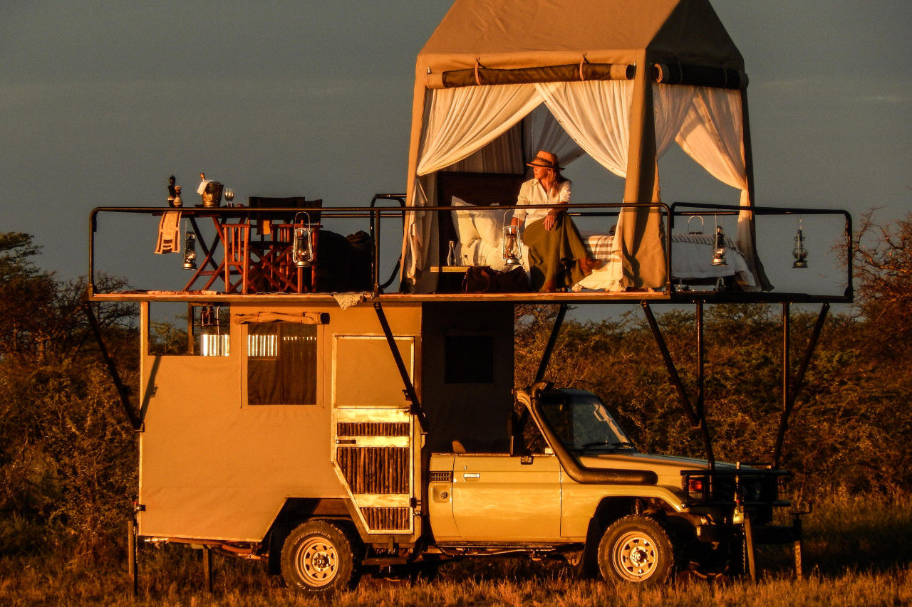 Frau sitzt auf einem luxuriösen Zeltbett auf einem Safari-Fahrzeug bei Sonnenuntergang in Afrika