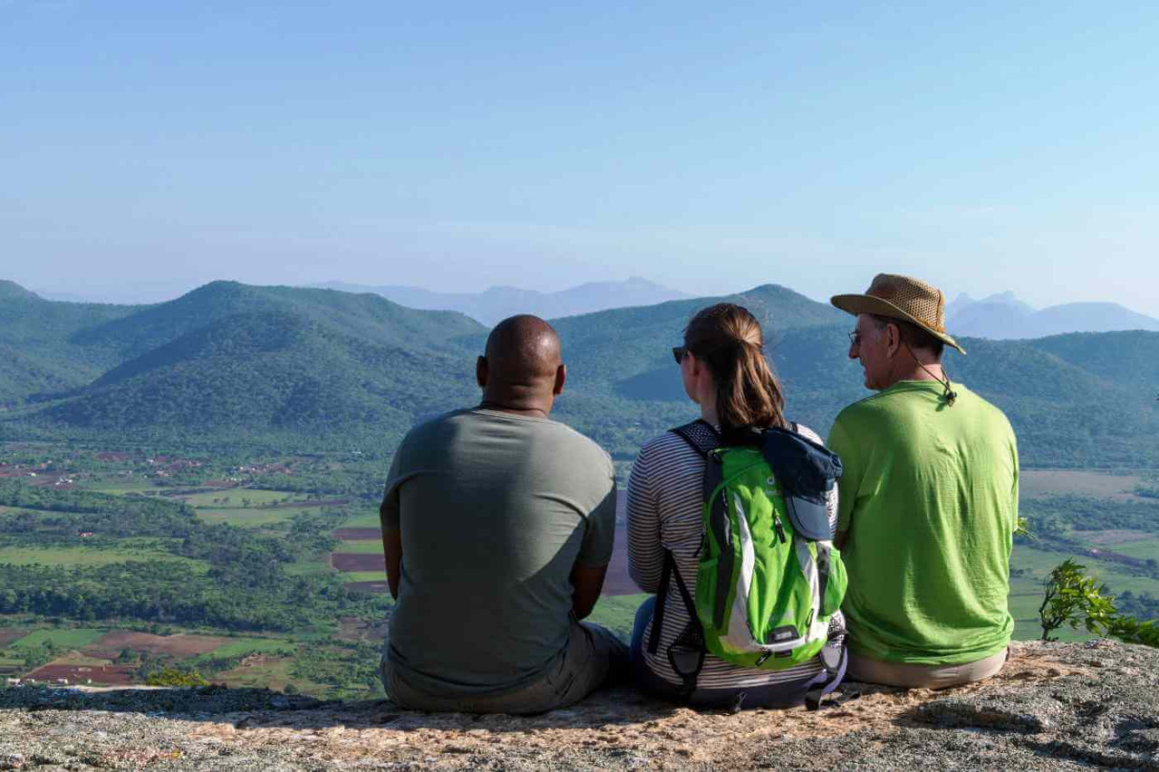 Drei Wanderer sitzen auf einem Felsen mit Panoramablick über grüne Hügel und Täler in Afrika
